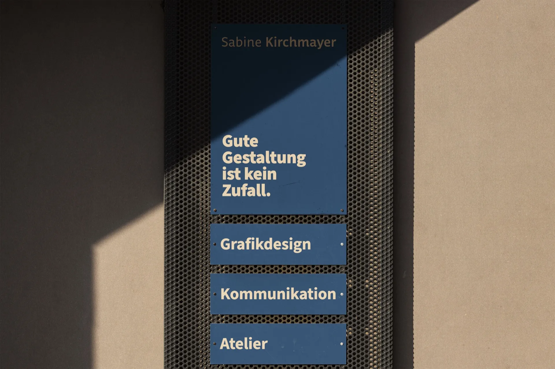 Hausschild an einer Fassade: großes blaues Schild mit Namenszug und Statement, darunter drei Zeilenschilder