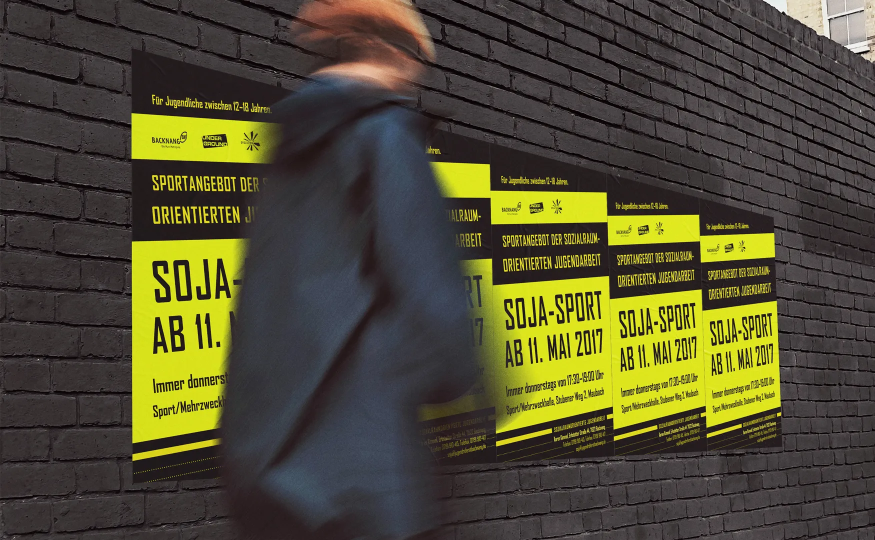 Gelbe Soja-Sport-Plakate an einer dunklen Ziegelwand, davor läuft eine Person vorbei