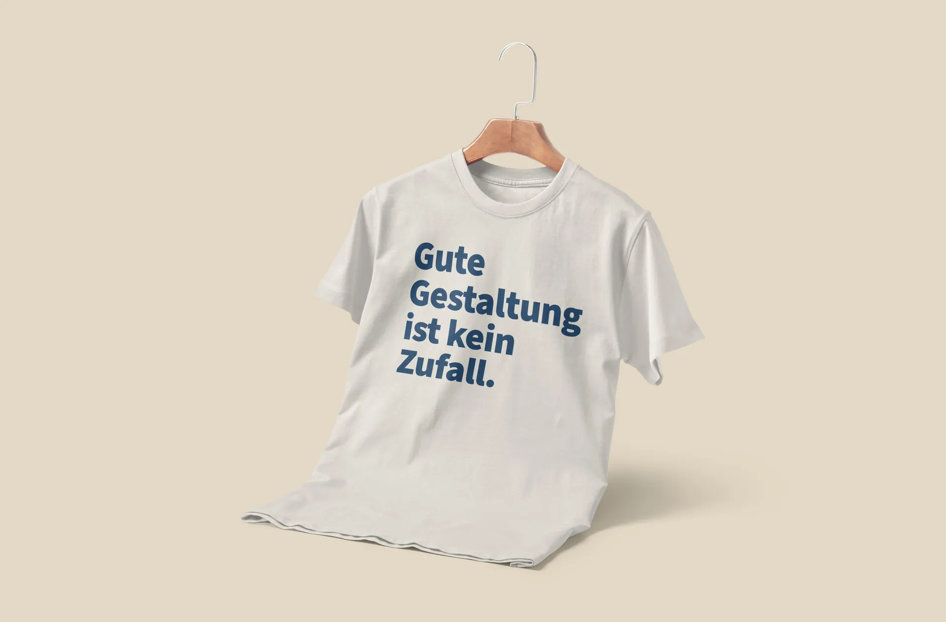 Cremefarbenes T-Shirt am Bügel mit blauem Brustdruck „Gute Gestaltung ist kein Zufall.“ auf beigem Hintergrund