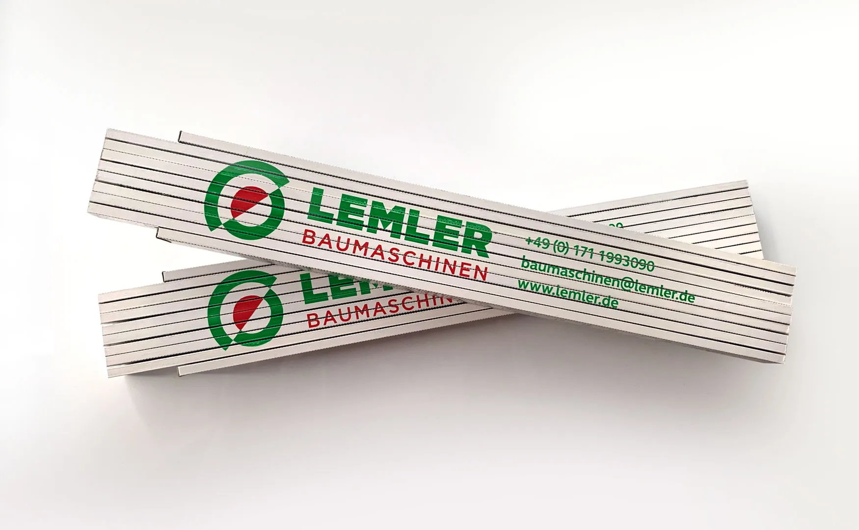 Zwei weiße Zollstöcke mit Logo und Kontaktdaten von Lemler Baumaschinen bedruckt